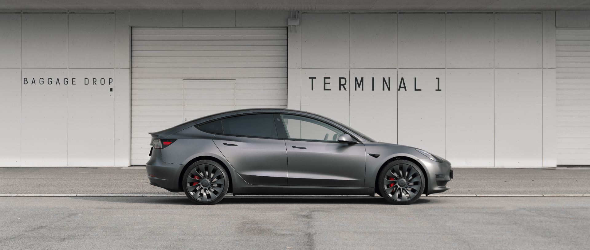 Grå Tesla Model Y set i profil foran en moderne lufthavnterminal — symbol på Drive Ons seriøse og retvisende vurdering af din bil